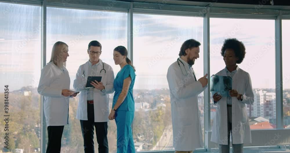 Mixed-races male and female professional doctors standing in modern clinic speaking and discussing Xray scan and typing on tablet. Group of healthcare workers in hospital at work. Medic concept Mixed-races male and female professional doctors standing in modern clinic speaking and discussing Xray scan and typing on tablet. Group of healthcare workers in hospital at work. Medic concept