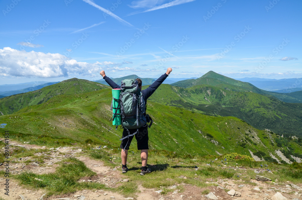Hiker with backpack standing on top of the mountain with raised hands in celebration of having reached goal during hiking travel trek
