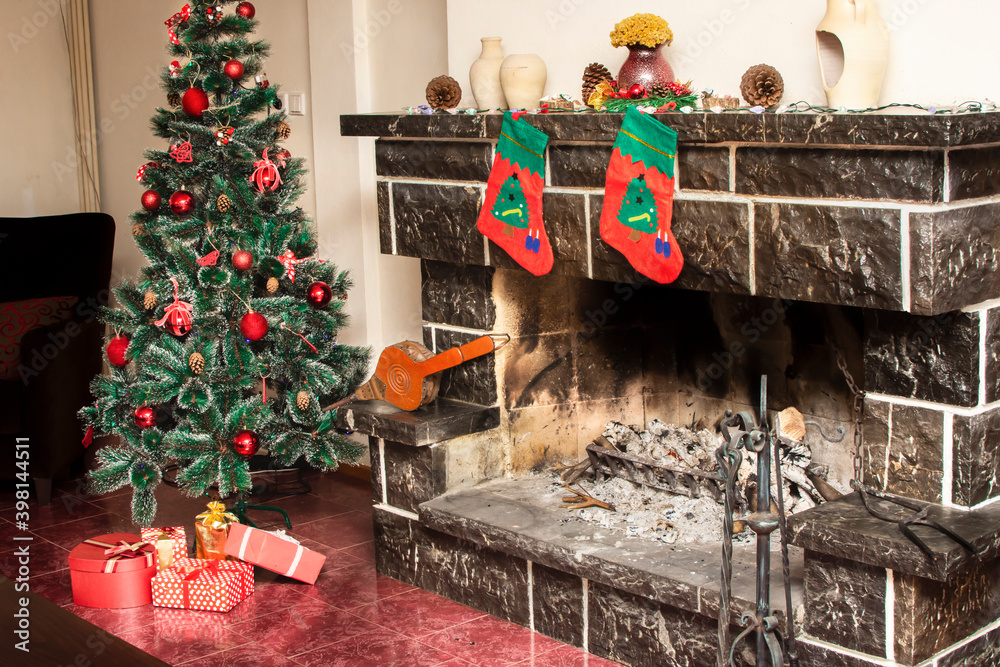 Fototapeta premium Christmas tree with presents near cozy fireplace with christmas socks in country house. New Year interior and home decoration. Winter or autumn vacation. Christmas holidays concept.