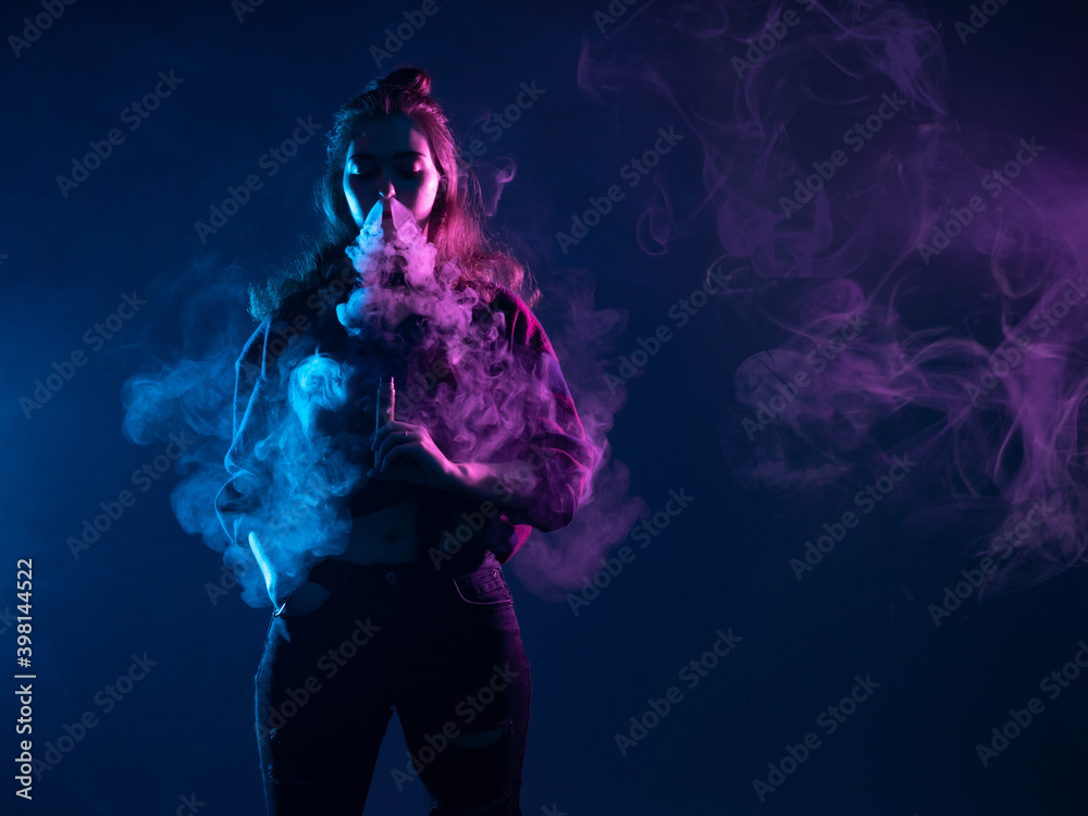 Vape girl releases steam from her nose. She is holding a vape device in her hand. portrait of vape girl standing in dark. Smoking electronic cigarettes. Woman vaper in cigarette smoke clubs.