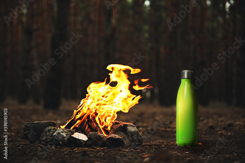 Green thermos bottle near bonfire with burning firewood in forest