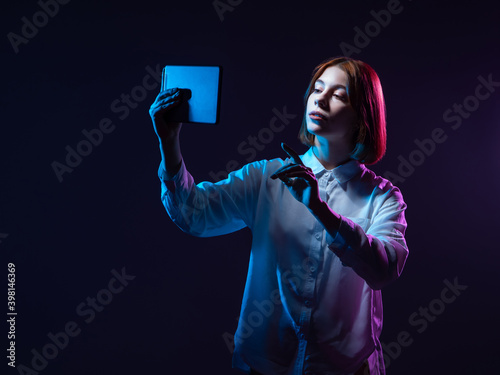 Fototapeta Naklejka Na Ścianę i Meble -  Woman with tablet in neon light. She lifts the tablet computer up. Girl on a dark background holds out a gadget. Tablet computer as a symbol of technology. Concept - videoconferencing technology.