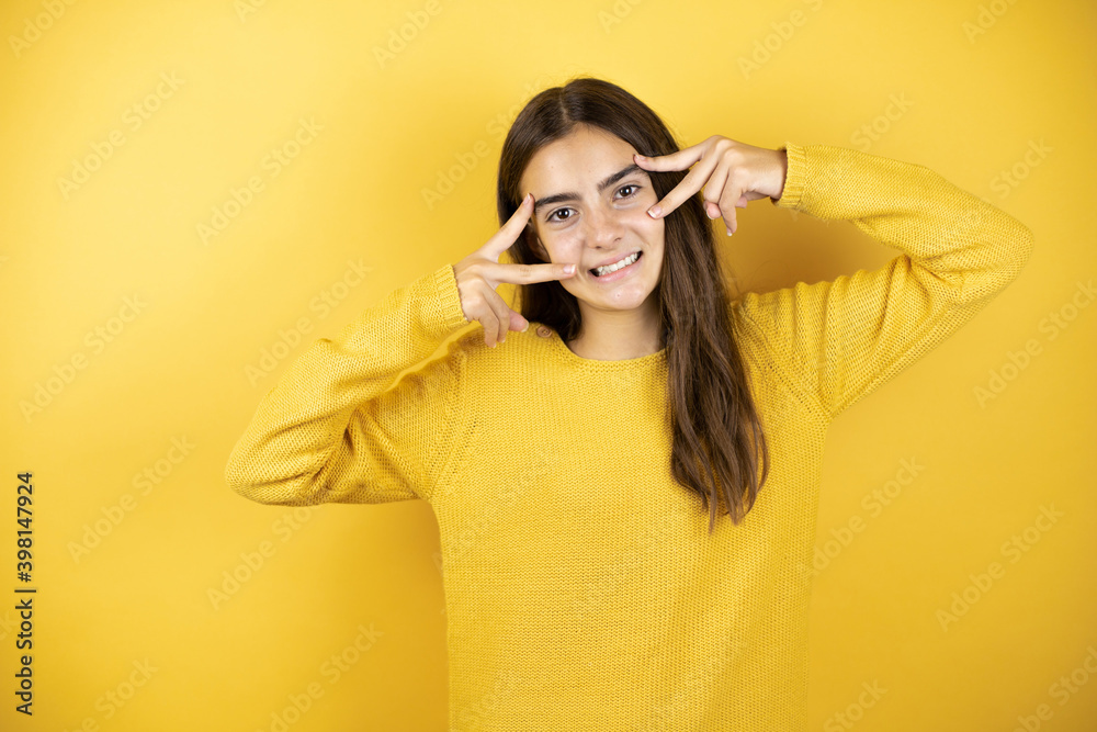Pretty girl wearing a yellow sweater standing over isolated yellow background Doing peace symbol with fingers over face, smiling cheerful showing victory Pretty girl wearing a yellow sweater standing over isolated yellow background Doing peace symbol with fingers over face, smiling cheerful showing victory