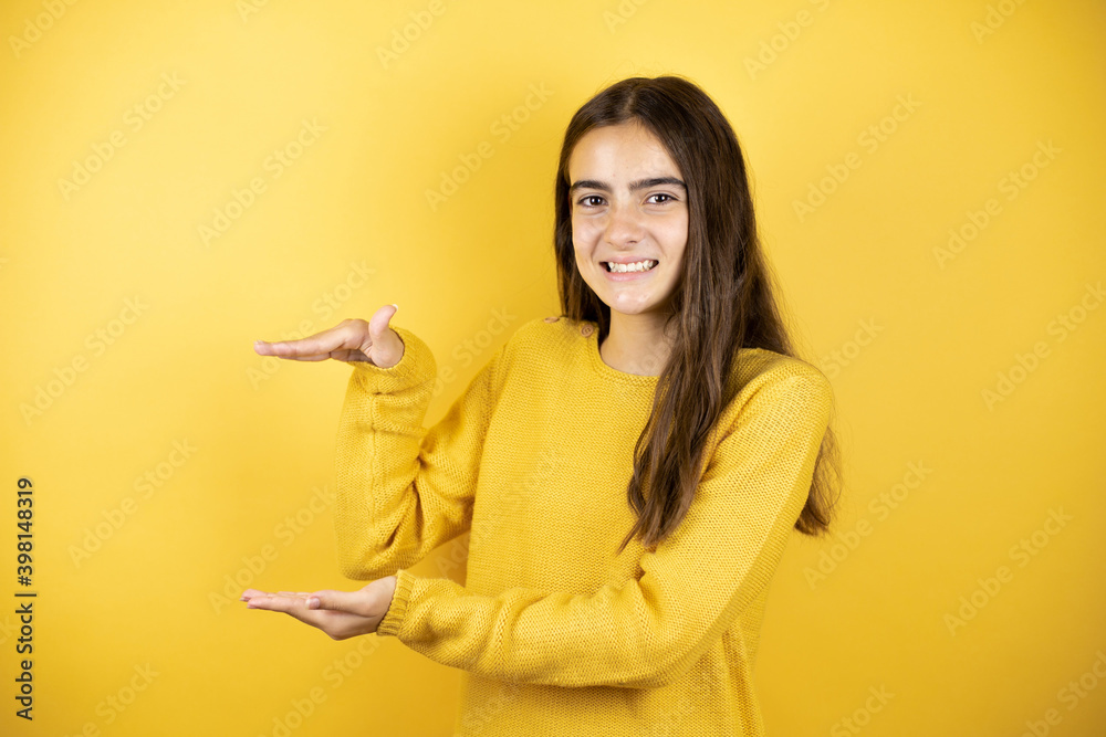 Pretty girl wearing a yellow sweater standing over isolated yellow background gesturing with hands showing big and large size sign, measure symbol. Smiling looking at the camera. Measuring concept. Pretty girl wearing a yellow sweater standing over isolated yellow background gesturing with hands showing big and large size sign, measure symbol. Smiling looking at the camera. Measuring concept.