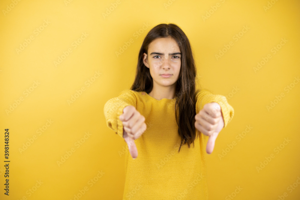Pretty girl wearing a yellow sweater standing over isolated yellow background with angry face, negative sign showing dislike with thumb down Pretty girl wearing a yellow sweater standing over isolated yellow background with angry face, negative sign showing dislike with thumb down