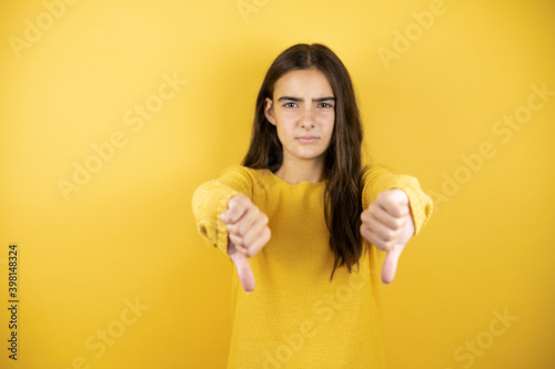Pretty girl wearing a yellow sweater standing over isolated yellow background with angry face, negative sign showing dislike with thumb down