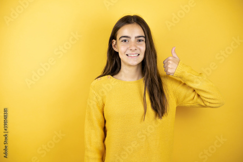 Pretty girl wearing a yellow sweater standing over isolated yellow background smiling confident doing the ok signal with her thumbs