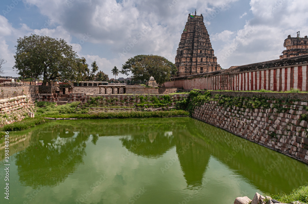 Hampi, Karnataka, India - November 4, 2013: Virupaksha Temple complex. Mamatha green water tank adjacent to outside wall under blue cloudscape. Reflection of East and central gate Gopurams.