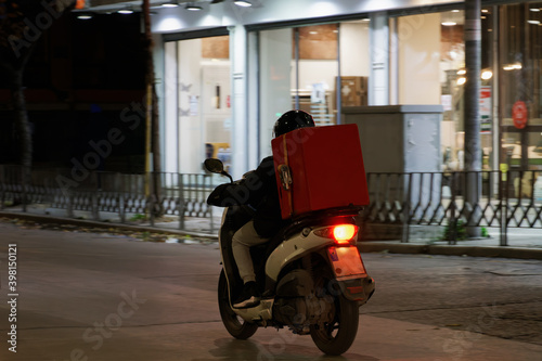 Delivery courier on unbranded motorbike speeding on city road at night. Illuminated view of male with helmet riding motorcycle to deliver take away food contained on a red box in Thessaloniki, Greece.