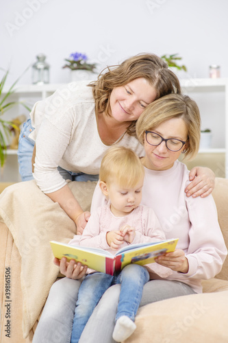 Two young women are sitting in the living room on the couch with a child on their knees and are reading a book to the child. Gay lesbian couple at home with a baby. Lesbian family concept. LGBT
