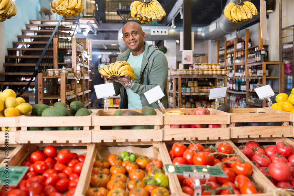 Positive Hispanic man choosing ripe sweet bananas in fruit and vegetable section of supermarket
