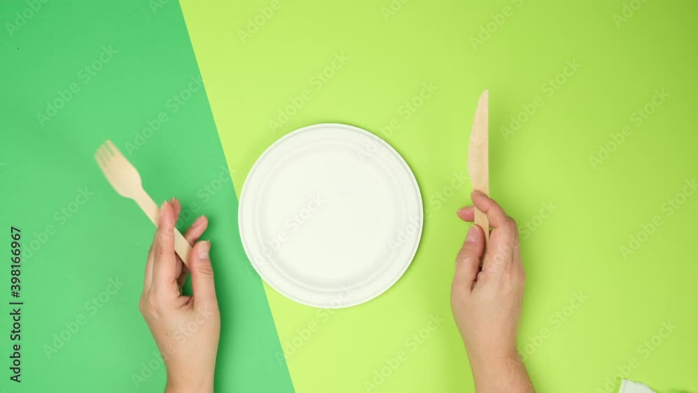 empty white paper plate and female hands are holding disposable fork and knife on a green background, top view