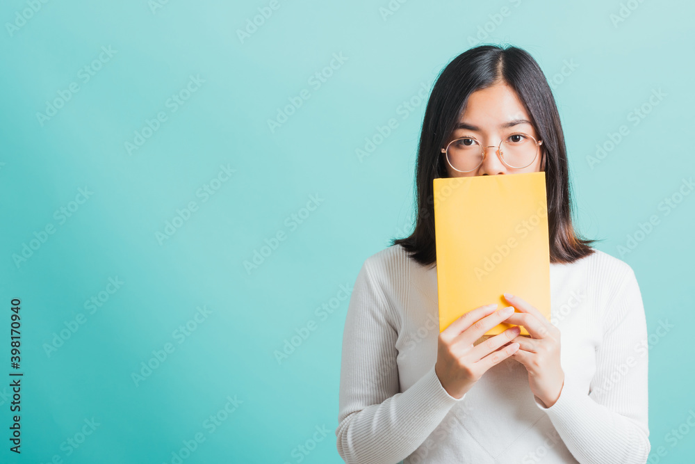 Young beautiful Asian woman hiding behind an open book, Portrait female in glasses is holding and reading a book, studio shot isolated on a blue background, Education concept Young beautiful Asian woman hiding behind an open book, Portrait female in glasses is holding and reading a book, studio shot isolated on a blue background, Education concept