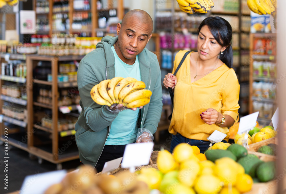 Obraz premium Smiling latin american family couple looking for ripe sweet bananas in food store..