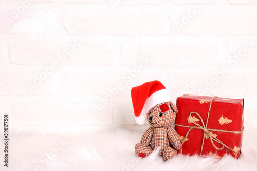 A little toy bear in a santa hat with a gift sits on the fur against the white brick wall.
