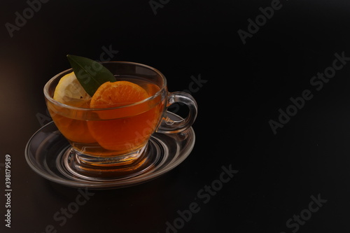 Liquid tea lemon orange slice green leaf cinnamon stick in transparent glass teacup saucer on black background