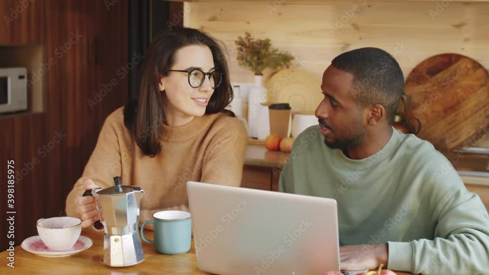 Young Caucasian woman sitting at kitchen table, pouring coffee into cups and discussing something on laptop with Afro-American husband Young Caucasian woman sitting at kitchen table, pouring coffee into cups and discussing something on laptop with Afro-American husband
