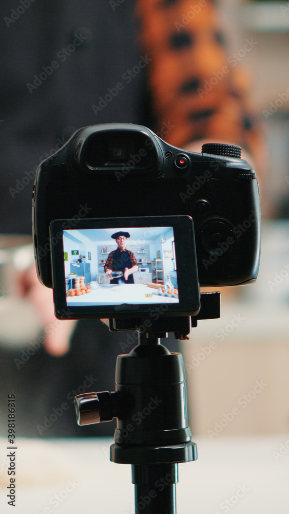 Close up of a video camera filming senior smiling man blogger in kitchen cooking. Retired blogger chef influencer using internet technology communicating on social media with digital equipment Close up of a video camera filming senior smiling man blogger in kitchen cooking. Retired blogger chef influencer using internet technology communicating on social media with digital equipment
