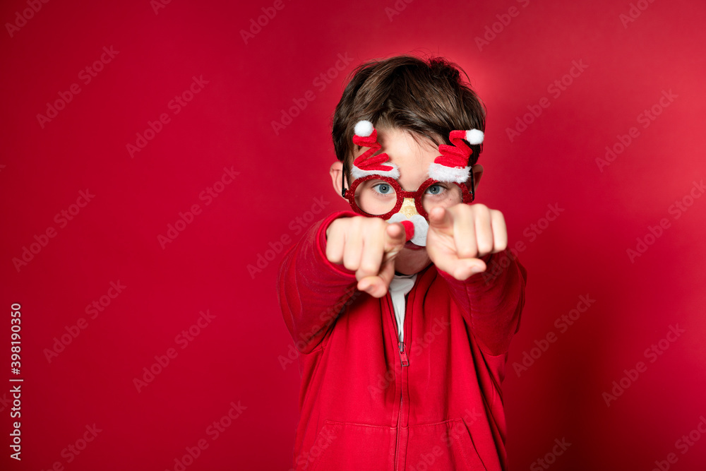 Obraz premium Pretty boy celebrating Christmas wearing funny Christmas glasses in front of red background