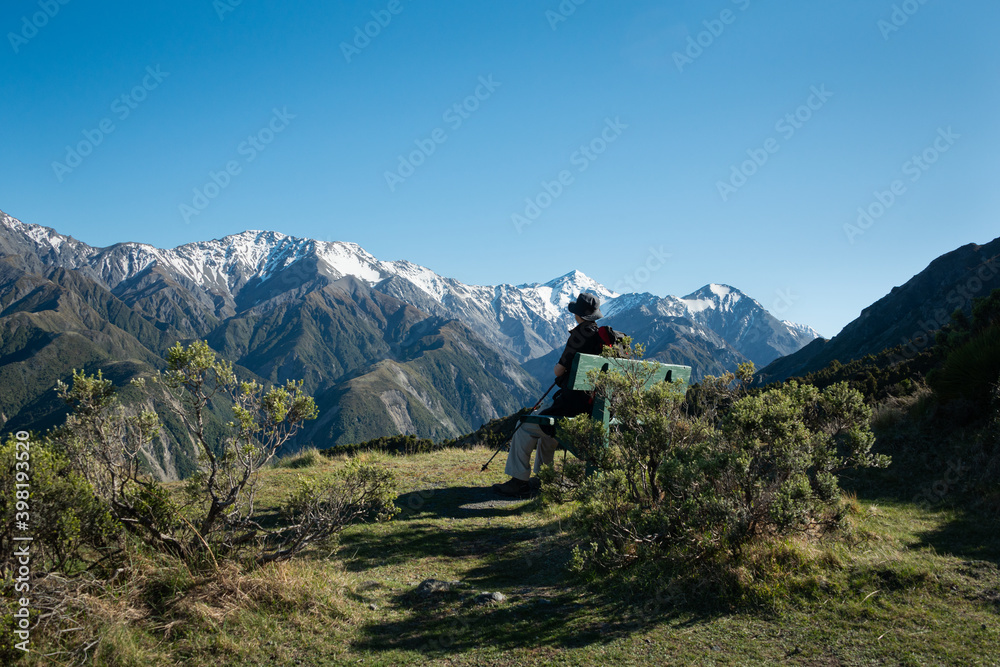 A backpacker sitting on the bench and admiring the snow-capped mountain peaks from Mt Fyffe track, South Island, New Zealand