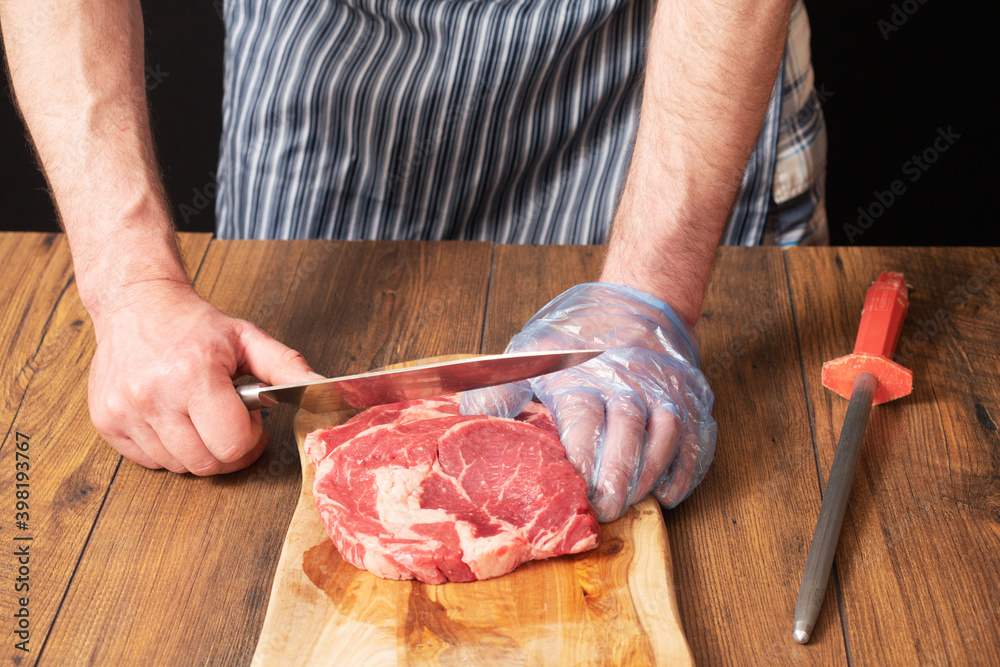 Professional butcher with knife and two rib eye steaks on a wooden cutting board. Butcher wears black and white stripe apron and blue gloves. Meat industry concept.