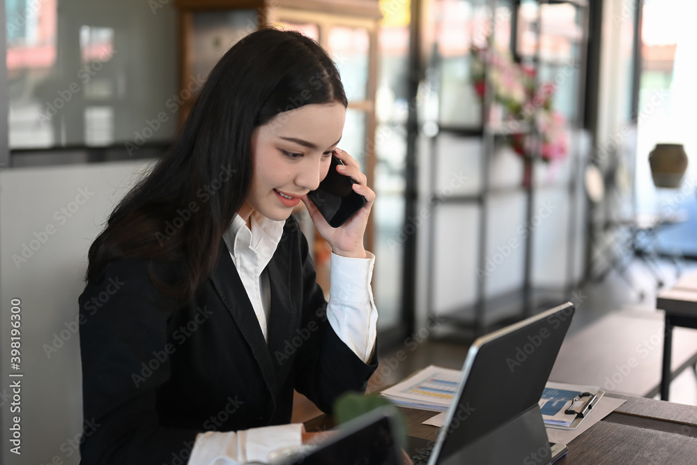 A young beautiful woman in black suit talking on mobile phone and using tablet while sitting at the office room. A young beautiful woman in black suit talking on mobile phone and using tablet while sitting at the office room.