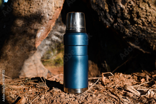 Thermos standing in a sunny forest. Vacuum travel flask close up. Camping concept, equipment for travel, adventure.