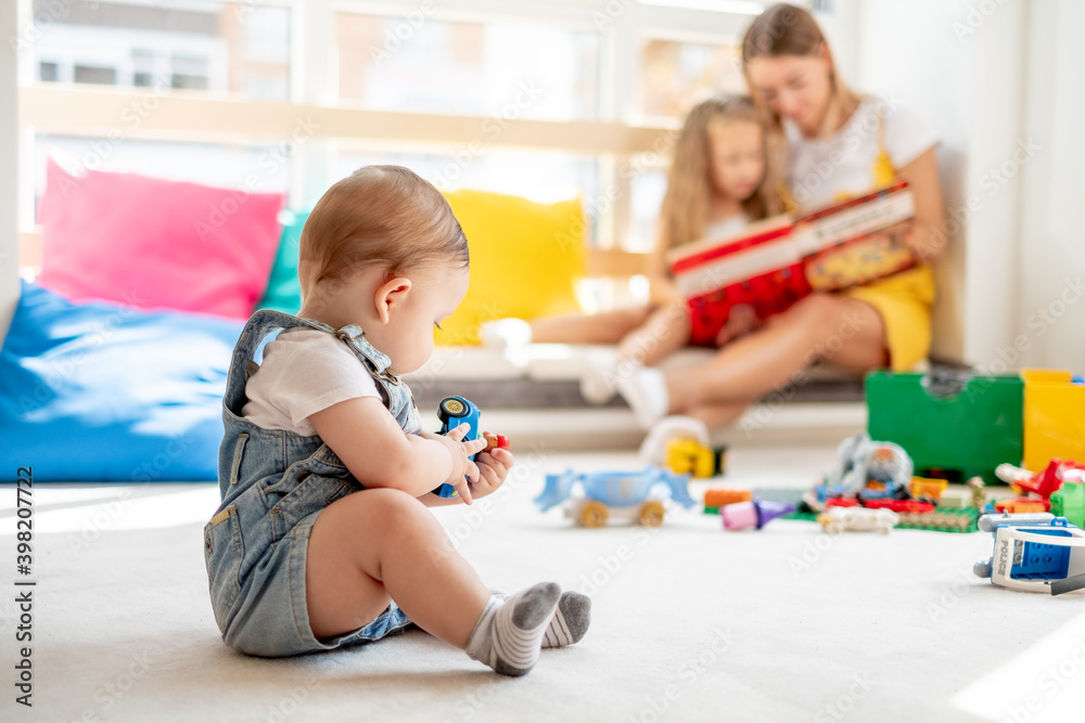 Baby playing with toys while mother and daughter reading book on background, family time, education Baby playing with toys while mother and daughter reading book on background, family time, education