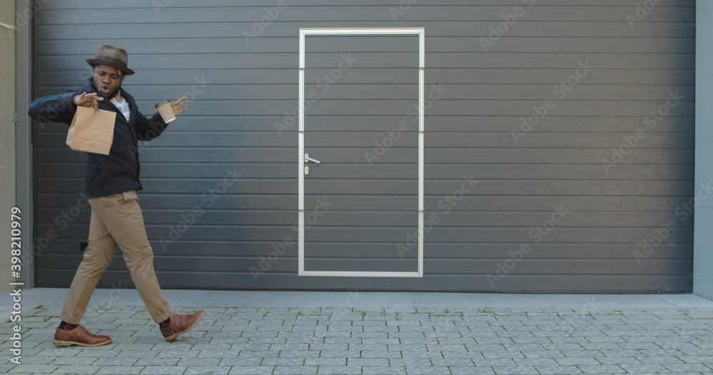 Stylish joyful man holding coffee paper cup and paper bag while moving in rhytm. Handsome afro american male person in hat dancing while walking near grey wall with doors Stylish joyful man holding coffee paper cup and paper bag while moving in rhytm. Handsome afro american male person in hat dancing while walking near grey wall with doors