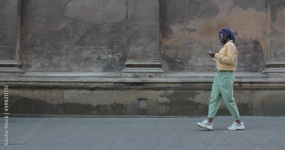 Joyful female person using smartphone and carrying coffee papper cup at city street. Afro american woman with dreadlocks walking and moving in rhytm. Concept of emotions