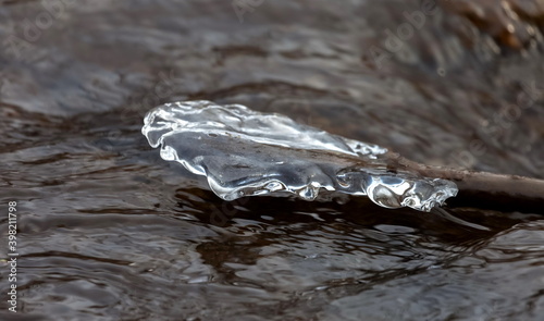 A piece of ice frozen on a tree branch close-up against the dark water of a fast river in early winter