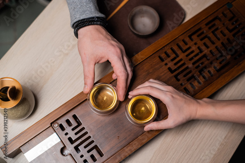 Top view of a bamboo board with a tray. Men's and women's hands close-up holding glass cups with amber tea at an intimate tea ceremony