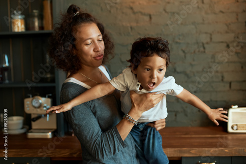 Portrait of carefree young mixed race female playing with her 2 year old baby son  holding him. Toddler boy pretending to fly like airplane in mother s hands. Maternity  happy childhood and fun