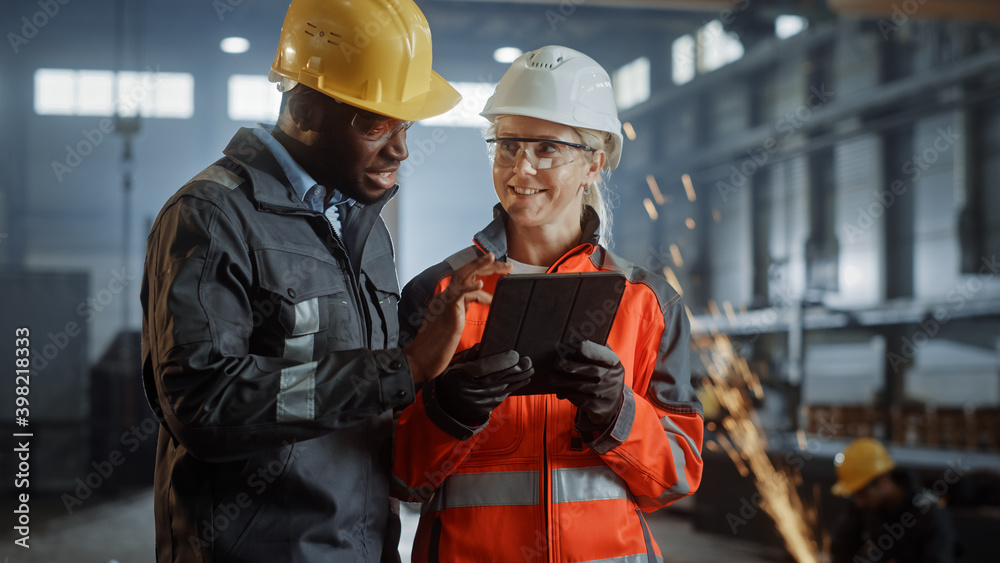 Two Heavy Industry Engineers Stand in Steel Metal Manufacturing Factory, Use Digital Tablet Computer and Have a Discussion. Black African American Industrial Specialist Talk to Female Technician. Two Heavy Industry Engineers Stand in Steel Metal Manufacturing Factory, Use Digital Tablet Computer and Have a Discussion. Black African American Industrial Specialist Talk to Female Technician.