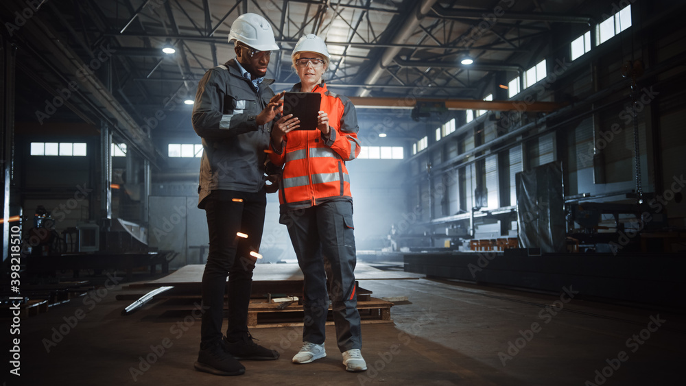 Fototapeta premium Two Heavy Industry Engineers Stand in Steel Metal Manufacturing Factory, Use Digital Tablet Computer and Have a Discussion. Black African American Industrial Specialist Talk to Female Technician.