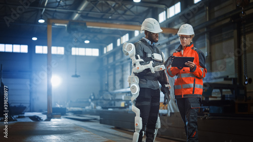 Futuristic Concept of a Manual Labor Worker in a Bionic Exoskeleton Prototype Working in a Factory. Heavy Industry Engineer Monitors the Powered Suit on an African American Assistant. photo