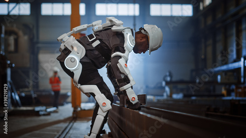Black African American Engineer is Testing a Futuristic Bionic Exoskeleton and Picking Up Metal Objects in a Heavy Steel Industry Factory. Contractor is Heavy Lifting Steel Parts in a Powered Shell. photo
