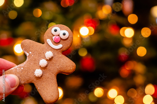 Hand holding a smiling gingerbread man in front of a blurred background with warm bokeh lights. Christmas lights background. Celebrating winter holidays, advent, Christmas season. Festive decoration.