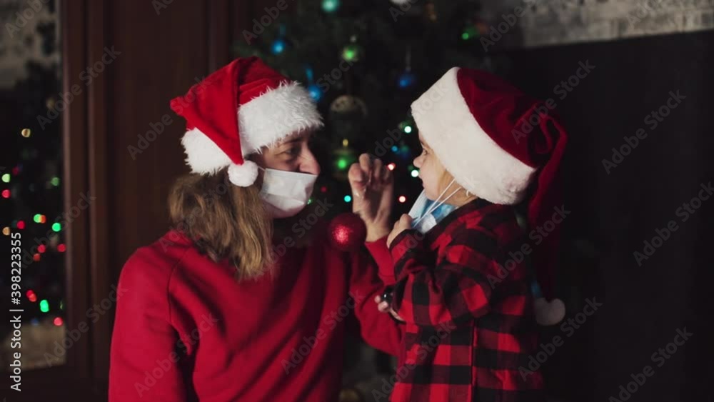 coronavirus, covid-19 in the new year. little daughter with her mother in a bad Santa hat and medical mask under the Christmas tree, remove the masks coronavirus, covid-19 in the new year. little daughter with her mother in a bad Santa hat and medical mask under the Christmas tree, remove the masks