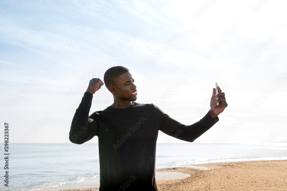 Young black sportsman on the beach who shows his strength while making a video call with his cell phone. Young black sportsman on the beach who shows his strength while making a video call with his cell phone.