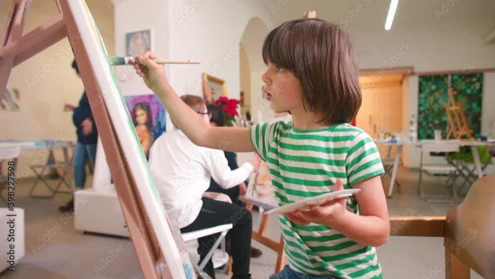 The creative atmosphere of an art studio. A cute long-haired boy of elementary school age paints a picture with a brush. Talent identification and development in children.