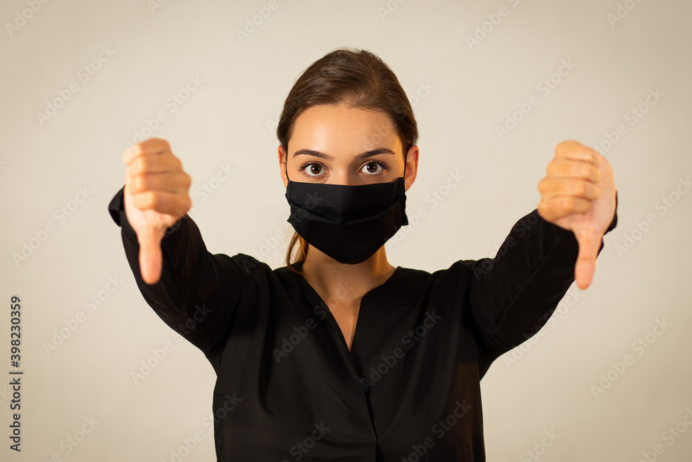 Unhappy young woman wearing black cloth face mask expressing her negative emotions. Girl in dark shirt with covered mouth and nose showing thumbs down. Unhappy young woman wearing black cloth face mask expressing her negative emotions. Girl in dark shirt with covered mouth and nose showing thumbs down.