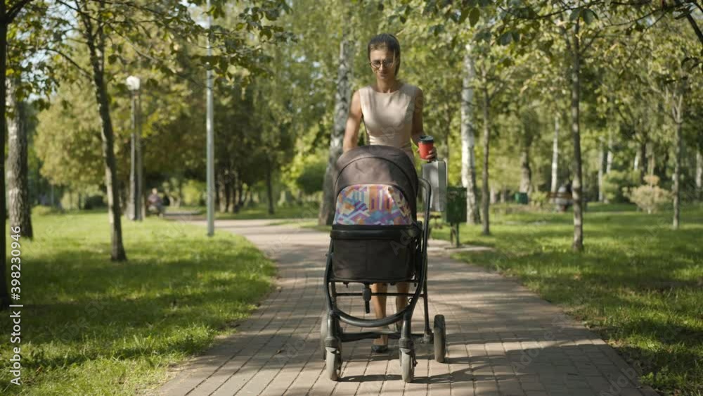 Wide shot of confident elegant businesswoman strolling in summer park with baby carriage. Positive Caucasian beautiful slim woman in eyeglasses walking outdoors on sunny day and smiling. Femininity. Wide shot of confident elegant businesswoman strolling in summer park with baby carriage. Positive Caucasian beautiful slim woman in eyeglasses walking outdoors on sunny day and smiling. Femininity.