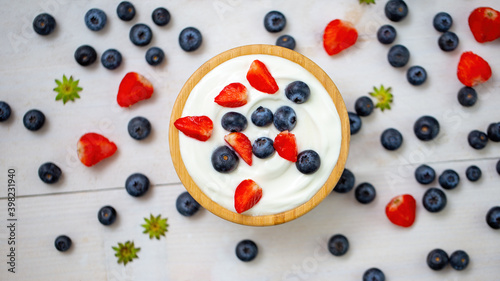 Bowl full of strawberries and blueberries with yogurth from directly above. Fresh healthy berries with dairy cream in wooden dish. Brown tray with little colorful fruits in background.