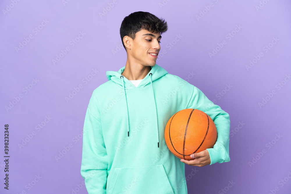 Young man playing basketball over isolated purple background looking side
