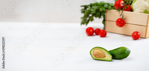 Fresh avocado on white table with space for text. Wooden box full of ripe tasty vegetable blurred in background. Green soft vitamin rich fruit cut in half in focus on desk. photo