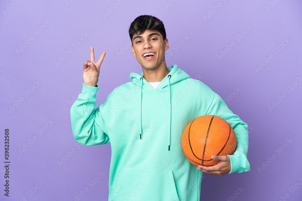 Young man playing basketball over isolated purple background smiling and showing victory sign