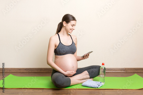 Pregnant female holding smartphone while sitting on exercise yoga mat at home at coronavirus time