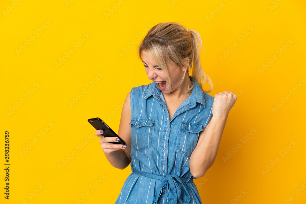Young Russian woman isolated on yellow background using mobile phone and doing victory gesture