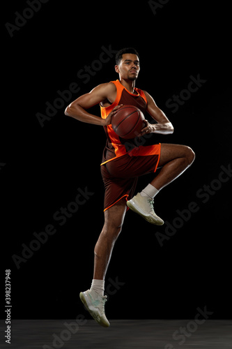 Jumping high. Young purposeful african-amrican basketball player training, practicing in action, motion isolated on black background. Concept of sport, movement, energy and dynamic, healthy lifestyle.